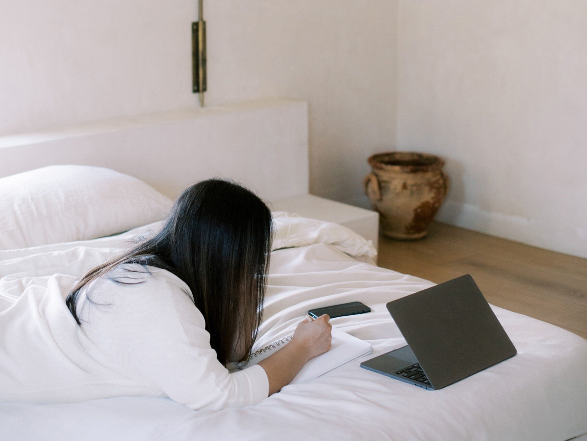 a woman lying on the bed writing on a notebook with an open laptop on her side